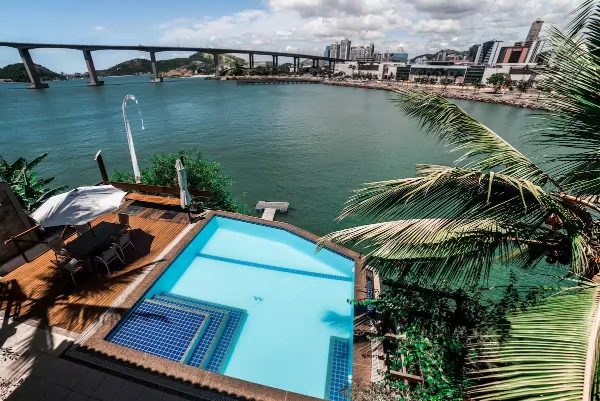 A casa vista do alto: piscina de borda infinita e deck na beira da baía, coqueiros e a Terceira Ponte ao fundo