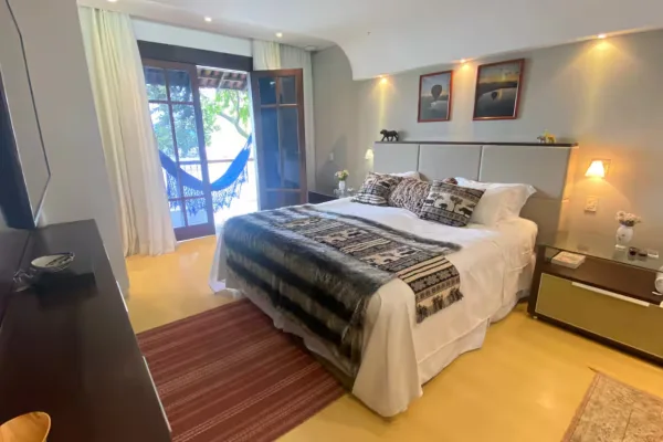 Suíte clara com cama de casal, tecidos africanos na colcha e a porta aberta para a varanda com rede