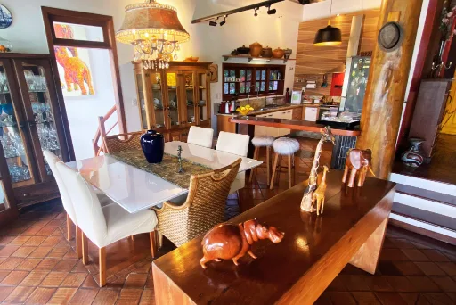 Sala de jantar com mesa branca, cristaleiras de madeira e a cozinha integrada ao fundo; na mesa de madeira da frente, girafas, elefante e hipopótamo entalhados